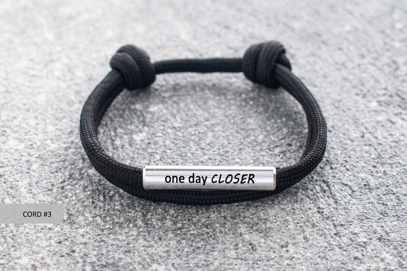 Op de afbeelding: Zwart paracord armband met een zilveren metalen balk waarop "one day CLOSER" staat.