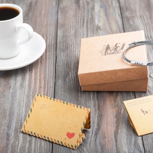Op de afbeelding: Een bruine kartonnen doos met de tekst "A&J Studio" erop gedrukt. De doos bevat een grijs gevlochten armband met een zilveren bedel waarop "Emily" staat. Er is een kopje koffie, een wit schoteltje en twee blanco kaarten met het woord "aan :" op een van de kaarten.