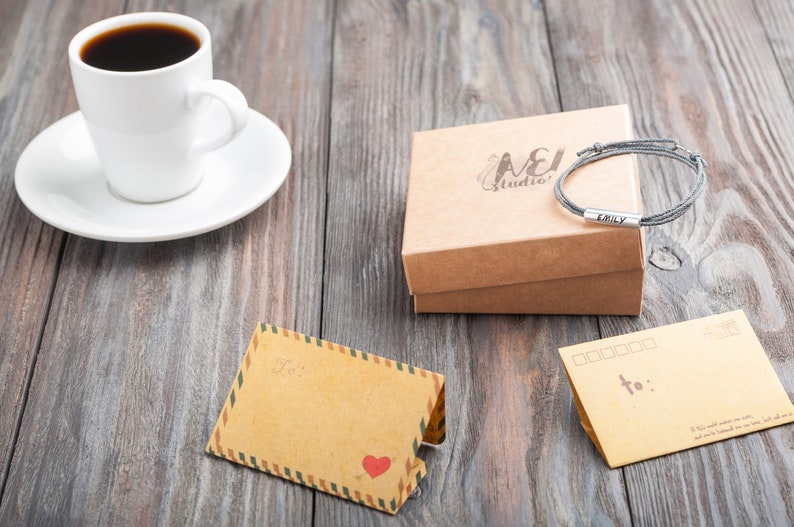 Op de afbeelding: Een bruine kartonnen doos met de tekst "A&J Studio" erop gedrukt. De doos bevat een grijs gevlochten armband met een zilveren bedel waarop "Emily" staat. Er is een kopje koffie, een wit schoteltje en twee blanco kaarten met het woord "aan :" op een van de kaarten.
