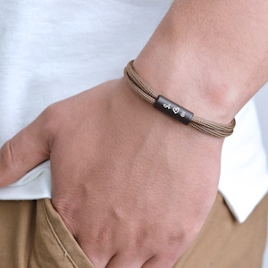 Pu&ograve; includere: Un braccialetto in paracord marrone con una barra in metallo nero incisa con le lettere "A &hearts; S".