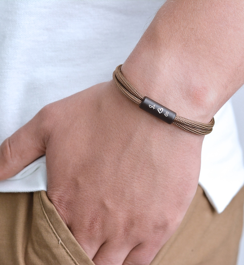 Pu&ograve; includere: Un braccialetto in paracord marrone con una barra in metallo nero incisa con le lettere "A &hearts; S".