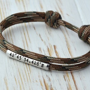 Op de afbeelding: Een bruin en groen camouflage paracord armband met een zilveren metalen tag gegraveerd met de co&ouml;rdinaten "42&deg; 43' 34.428" N"