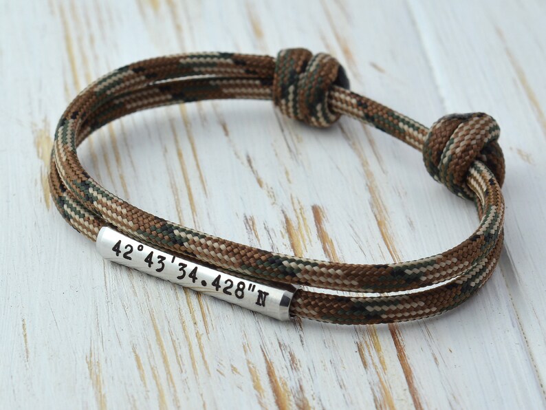 Op de afbeelding: Een bruin en groen camouflage paracord armband met een zilveren metalen tag gegraveerd met de co&ouml;rdinaten "42&deg; 43' 34.428" N"