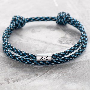 Op de afbeelding: Een gevlochten armband van zwart, wit en blauw touw met een zilveren bedel met de tekst "K.C."