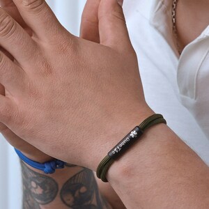 Puede incluir: Una pulsera de paracord verde con un dije de metal negro que dice "Diab&eacute;tico" y un s&iacute;mbolo m&eacute;dico.