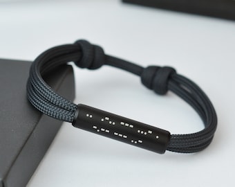 May include: Black bracelet with a braided cord design. The bracelet features a cylindrical black metal accent with white Morse code dots and dashes. The cord is knotted at both ends for adjustment.