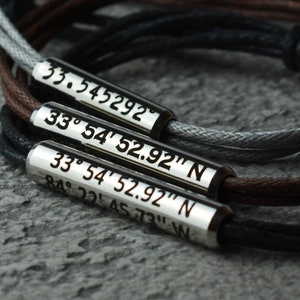 May include: A set of three silver bracelets with black, brown, and gray cords. Each bracelet has a silver tag with coordinates engraved on it. The coordinates are 33.545292, 33&deg; 54' 52.92" N, and 33&deg; 54' 52.92" N 84&deg; 22&prime; 4573" W.