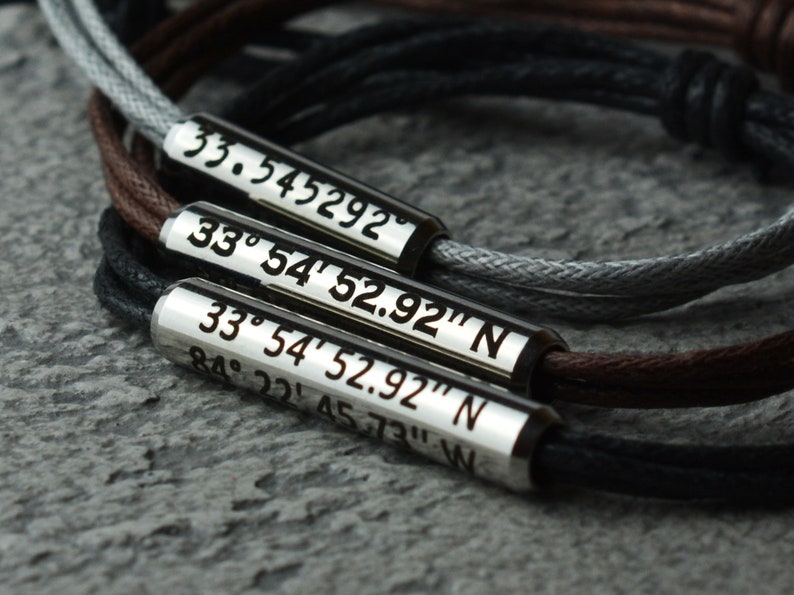 May include: A set of three silver bracelets with black, brown, and gray cords. Each bracelet has a silver tag with coordinates engraved on it. The coordinates are 33.545292, 33&deg; 54' 52.92" N, and 33&deg; 54' 52.92" N 84&deg; 22&prime; 4573" W.