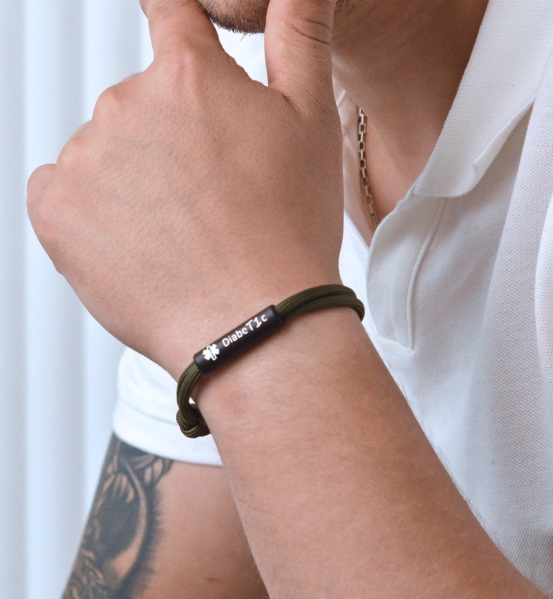 Puede incluir: Una pulsera de paracord verde con un dije de metal negro que dice "Diab&eacute;tico" y un s&iacute;mbolo de tr&eacute;bol de cuatro hojas.