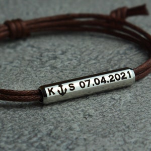 May include: Brown leather cord bracelet with a silver bar charm engraved with the letters K and S, an anchor symbol, and the date 07.04.2021.