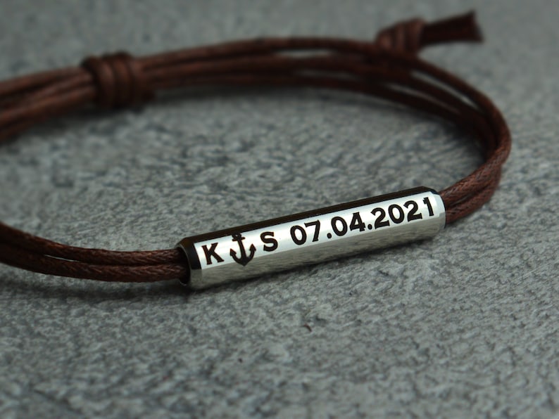 May include: Brown leather cord bracelet with a silver bar charm engraved with the letters K and S, an anchor symbol, and the date 07.04.2021.