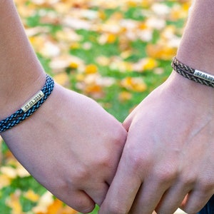 May include: Two matching friendship bracelets with black and blue or brown and green paracord. The bracelets have a silver charm with the date "07.01.2018" engraved on it.