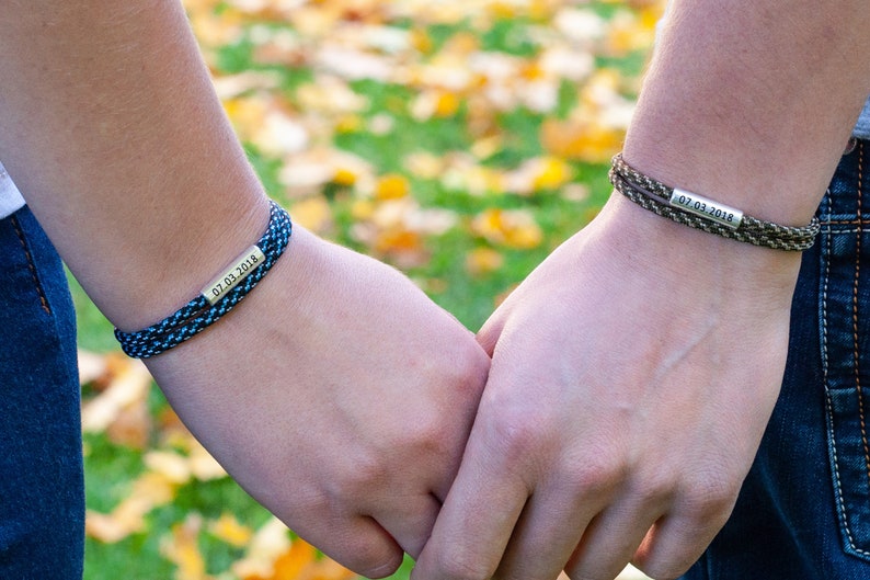 May include: Two matching friendship bracelets with black and blue or brown and green paracord. The bracelets have a silver charm with the date "07.01.2018" engraved on it.
