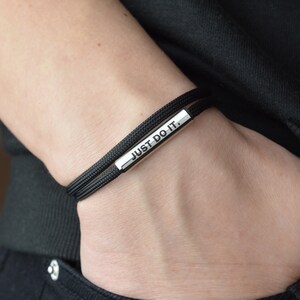Op de afbeelding: Zwarte armband met een zilveren balk waarop "JUST DO IT." staat.