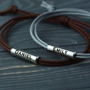 May include: Two personalized bracelets with brown and gray cords. The bracelets have metal tags with the names "Daniel" and "Emily" engraved on them.