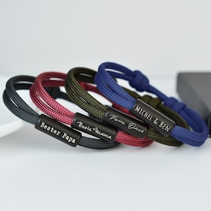 May include: Four different colored braided rope bracelets with personalized engraved metal plates. The bracelets are in the colors red, black, green, and blue. The plates read "Bester Papa", "Beste Mama", "Vivre Ouvre", and "Michel & Ben".
