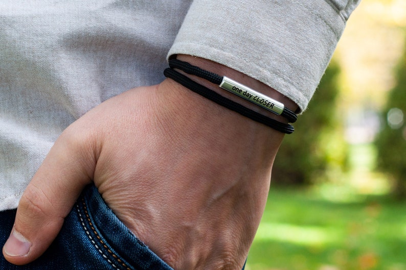 Op de afbeelding: Een zwarte paracord armband met een zilveren staafbedel met de tekst "one day closer".