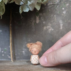 May include: A small wooden doll with brown hair and a white crocheted dress. The doll is standing on a wooden surface.
