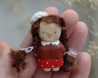 May include: A handmade wooden doll with brown hair, a white hat, and a red and white dress. The doll is wearing a brown jacket with a white collar.