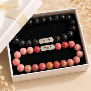 May include: Two beaded bracelets inside a white gift box. One bracelet features black porous lava beads with a silver-toned rectangular charm engraved with A & M. The second bracelet features smooth pink and orange stone beads with a silver-toned charm engraved with M & A.