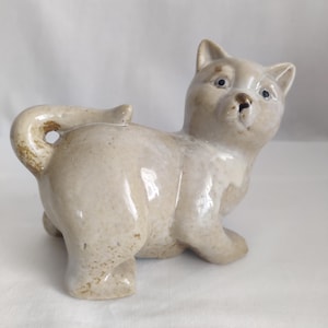 May include: A ceramic cat figurine with a white glaze and brown speckles. The cat is sitting with its tail curled around its body.