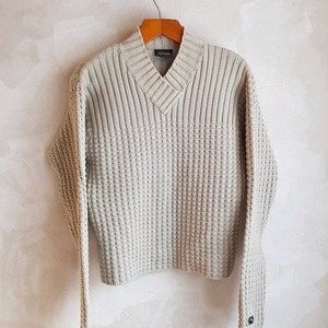 May include: A light grey knitted sweater with a v-neck and a waffle knit pattern. The sweater is hanging on a wooden hanger.