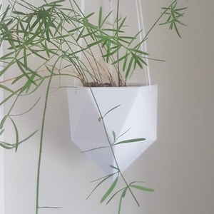 May include: A white geometric hanging planter with a green plant inside. The planter is made of a smooth, white material and has a diamond-shaped design.