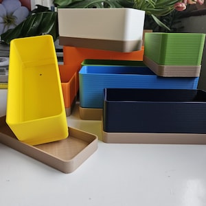 May include: A selection of rectangular planters in various colours, including yellow, orange, blue, green, and white. Each planter has a contrasting brown base. The planters are stacked and arranged on a white surface.