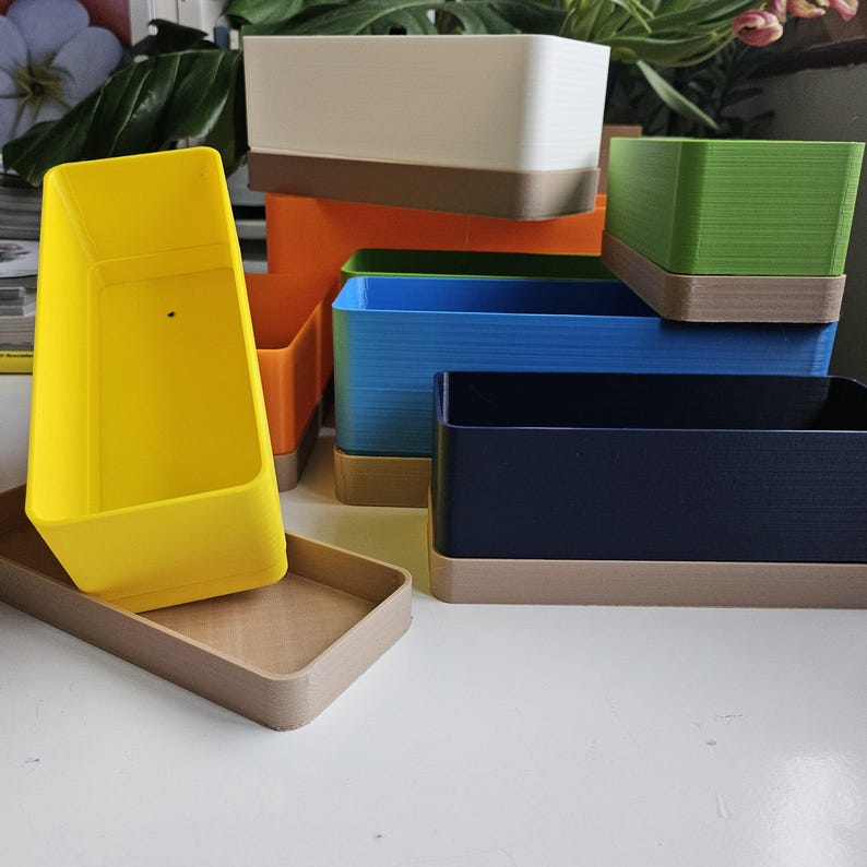 May include: A selection of rectangular planters in various colours, including yellow, orange, blue, green, and white. Each planter has a contrasting brown base. The planters are stacked and arranged on a white surface.