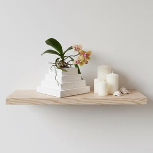 May include: A white stepped pyramid planter containing a phalaenopsis orchid with yellow and pink flowers, accompanied by three white pillar candles of varying heights and two small white seashells arranged on a light-toned wooden floating shelf.