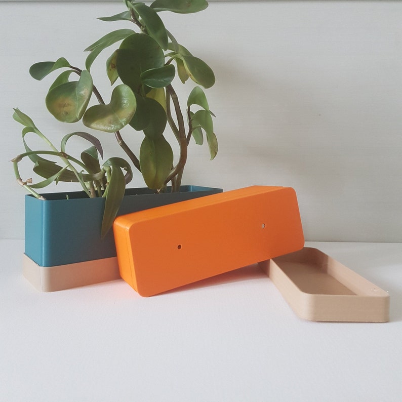 May include: A rectangular teal and beige planter with a vibrant green plant. An orange lid and a beige tray are next to the planter. The planter is a modern design, suitable for indoor or outdoor use.