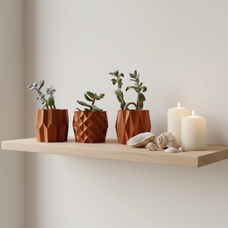 May include: Three terracotta-coloured geometric planters containing green succulents, arranged alongside two white pillar candles and three small seashells on a light-toned wooden shelf. The planters feature distinct faceted, textured surfaces.