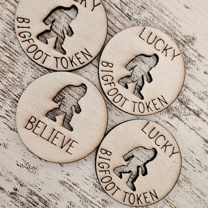 May include: Four wooden Bigfoot tokens. Each token is round and features a Bigfoot silhouette cutout. The tokens are engraved with the words "LUCKY" and "BIGFOOT TOKEN" around the edge, and one token says "BELIEVE".