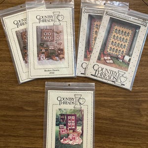 May include: Several Country Threads cross-stitch pattern packets. The packets are in clear plastic sleeves. The packets feature images of finished cross-stitch projects with titles like "Broken Hearts" and "Father Christmas."