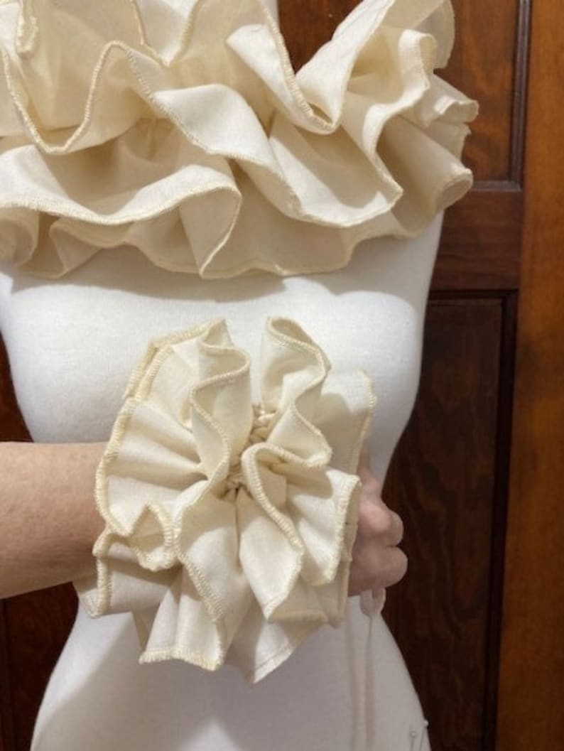 May include: A pair of off-white fabric ruffles, one large and one small. The large ruffle is a collar, and the small ruffle is a wrist cuff. Both ruffles are made of soft, gathered fabric with a delicate, scalloped edge.