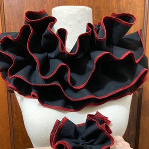 May include: A black fabric neck ruff featuring multiple layers of ruffled fabric with a vibrant red stitched edge. The accessory is displayed on a white mannequin form, accompanied by a smaller, matching black and red ruffled fabric piece.