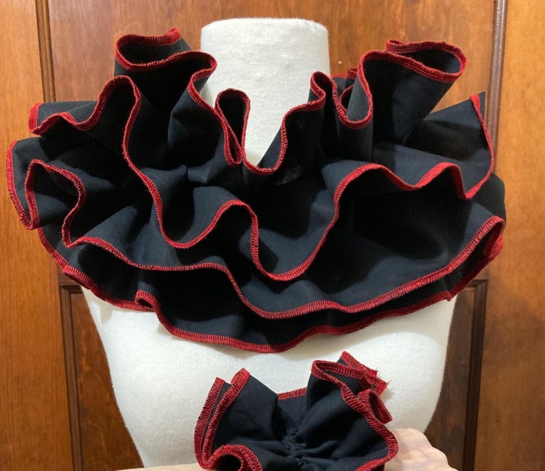 May include: A black fabric neck ruff featuring multiple layers of ruffled fabric with a vibrant red stitched edge. The accessory is displayed on a white mannequin form, accompanied by a smaller, matching black and red ruffled fabric piece.