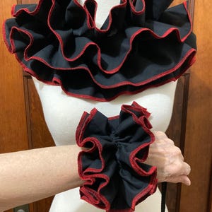 May include: A black fabric neck ruff and a matching wrist cuff, both featuring multiple layers of ruffled fabric with a contrasting red stitched edge. The neck ruff is wide and circular, while the wrist cuff is smaller and gathered.