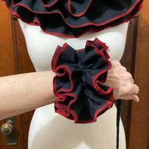May include: A black fabric wrist cuff featuring dense, layered ruffles with a contrasting red stitched edge. The cuff is designed with a gathered, voluminous texture that encircles the wrist, showcasing a dark color palette and detailed trim.