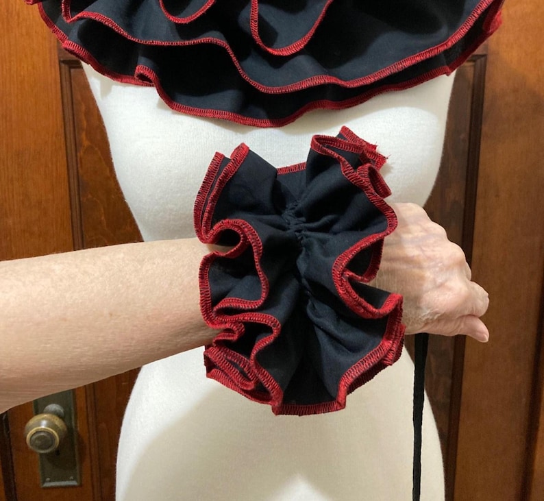 May include: A black fabric wrist cuff featuring dense, layered ruffles with a contrasting red stitched edge. The cuff is designed with a gathered, voluminous texture that encircles the wrist, showcasing a dark color palette and detailed trim.