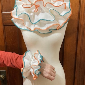 May include: White ruffled collar and matching cuffs with orange and teal trim. The collar is large and dramatic, while the cuffs are smaller and more delicate.