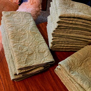 May include: Several stacks of sage green quilted fabric items are arranged on a dark wooden surface. The fabric has a repeating floral pattern. The items appear to be folded and stacked, suggesting they are blankets or bedspreads.