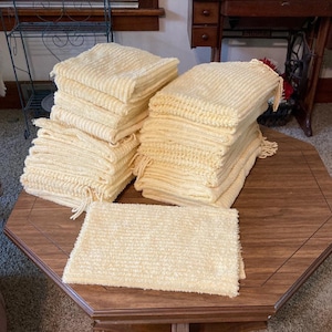May include: A collection of pale yellow rectangular towels with a textured, ribbed surface and fringed edges. The towels are arranged in two neat stacks and one single piece laid flat, showcasing a consistent light color and woven fabric pattern.