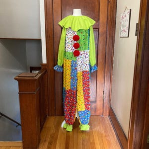 May include: A colorful clown costume with a lime green collar and sleeves, and a patchwork design. The costume features red pom-poms and ruffled cuffs and ankles. The outfit is displayed on a mannequin.