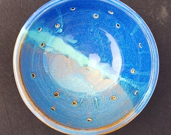 May include: A handmade ceramic bowl with a vibrant blue glaze. The bowl features a series of evenly spaced holes around the rim and a contrasting light blue and white interior. The rim has a brown border.