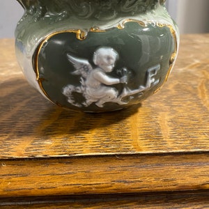 May include: A green ceramic planter with a white cherub design and gold accents. The cherub is holding a flower and a small bird.