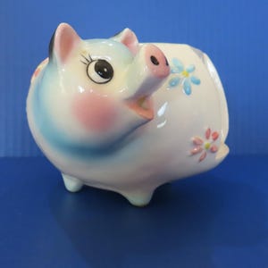 May include: A ceramic piggy bank in the shape of a pig, with a white body, blue and pink accents, and small floral designs. The pig has a smiling expression and is set against a blue background.