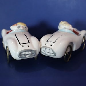 Könnte beinhalten: Ein Paar weiße Keramik-Salz- und Pfefferstreuer in Form von Rennwagen. Jeder Wagen hat einen Fahrer, schwarze Details und goldfarbene Räder. Die Streuer stehen auf einer dunkelblauen Oberfläche.