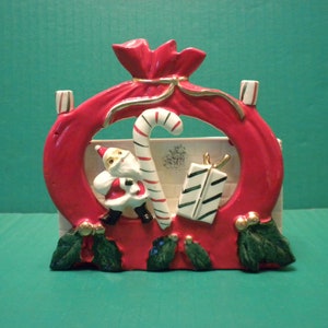 Papai Noel da Ucagco com um bastão de doces e presentes na frente de uma silhueta de saco de Papai Noel vermelho amarrado com uma fita dourada - com detalhes de azevinho - suporte para bastão de doces