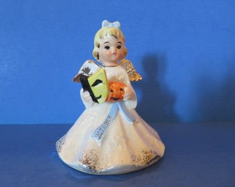 Könnte beinhalten: Eine Keramikfigur eines jungen Engels mit blondem Haar, goldenen Flügeln und einem weißen Kleid. Der Engel hält eine grüne und schwarze Maske und einen kleinen orangefarbenen Kürbis. Ein blaues Band mit dem Wort "OKTOBER" verläuft über das Kleid.